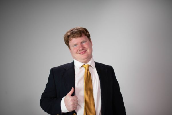 Studio headshot of Elliot Dennis in a navy suit and gold tie, giving a thumbs up.