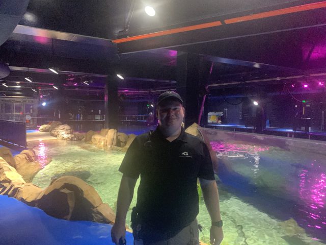 Elliot in a work polo and cap standing beside a brightly lit aquarium habitat pool at night, the gallery empty behind him.