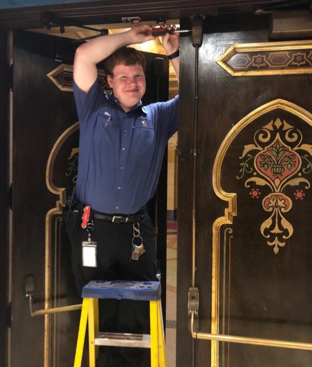 A younger Elliot on a step ladder adjusting a door closer above the ornate gilded doors of the Fox Theatre, keys and radio on his belt.