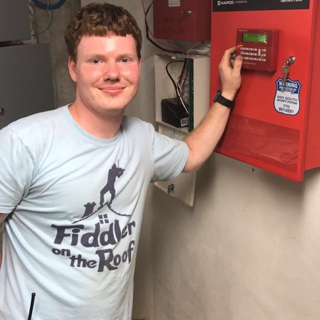 A teenage Elliot in a "Fiddler on the Roof" t-shirt with one hand on a red commercial fire alarm control panel, grinning at the camera.