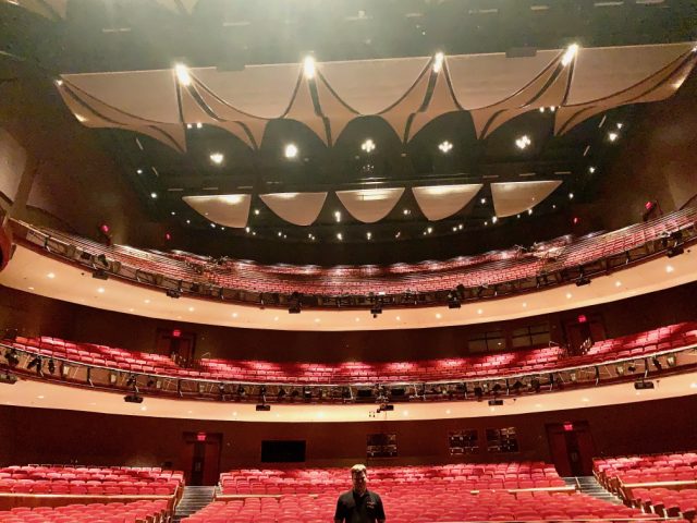 A large performing arts theatre photographed from the stage: three tiers of empty red seats and a sculpted acoustic canopy overhead, with Elliot standing alone in the middle of the house.
