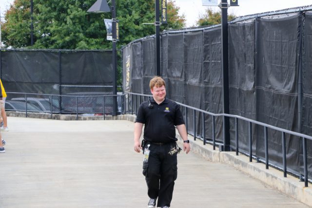 Elliot in a black KSU polo with a radio and tool pouch, walking a campus concourse past Owls banners on an event day.