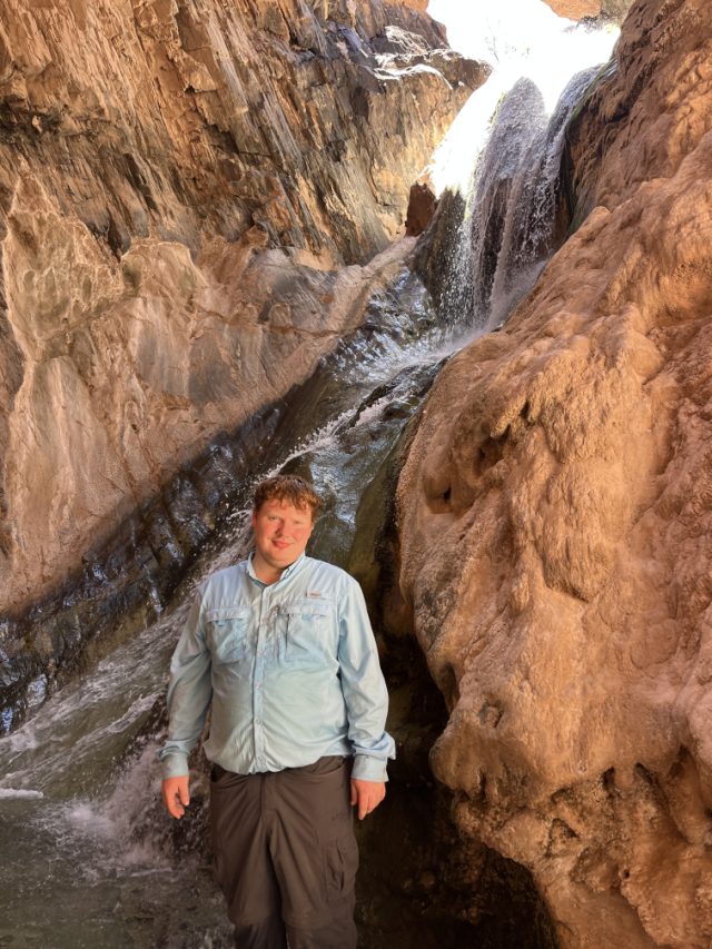 Elliot in a light blue hiking shirt standing in a narrow desert canyon, a waterfall running down travertine rock behind him.
