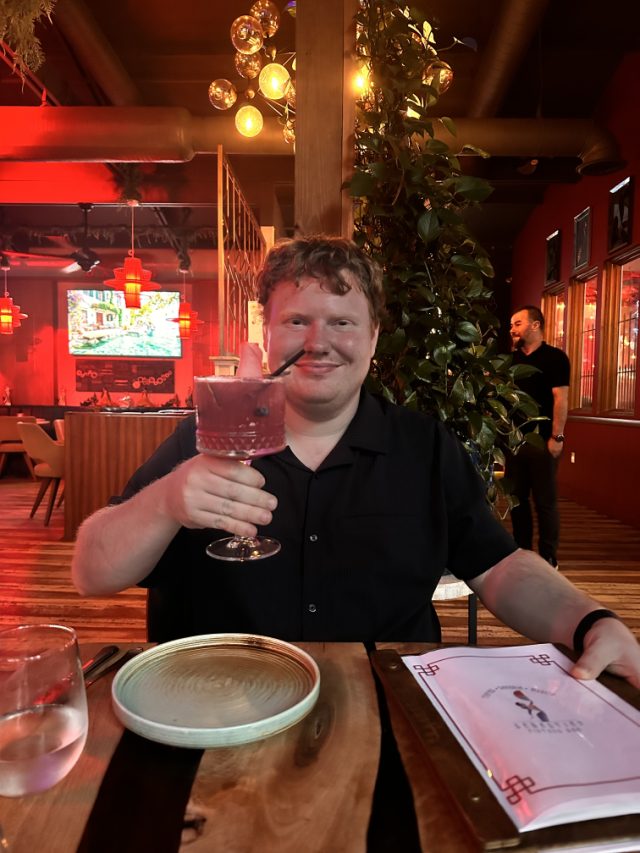 Elliot raising a pink cocktail in a dim restaurant lit red, lanterns and hanging greenery around him.
