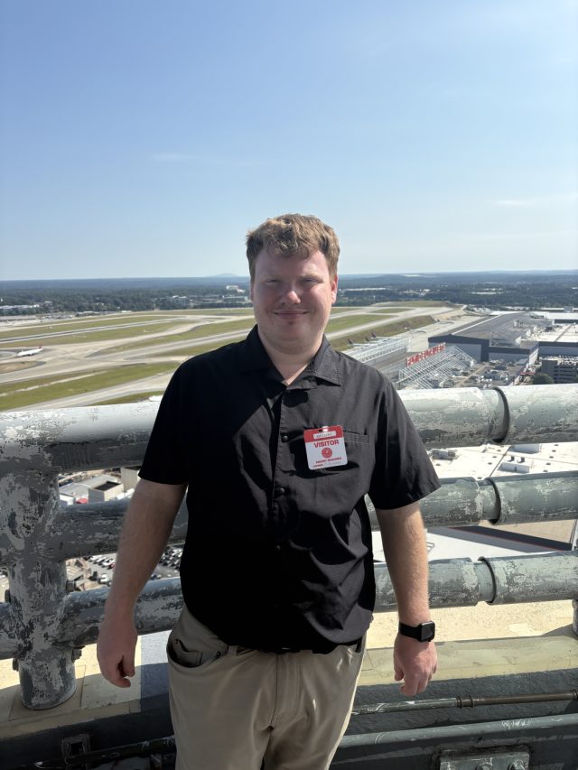 Elliot on an airport rooftop wearing a red visitor badge, with taxiways, runways and a maintenance hangar spread out behind him.