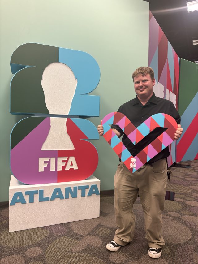 Elliot holding a large colourful heart-shaped prop beside a FIFA World Cup 26 Atlanta trophy sculpture.