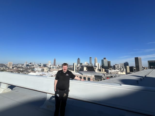 Elliot on the roof of Mercedes-Benz Stadium in a black work polo, tools and radio on his belt, the downtown Atlanta skyline spread out behind him.