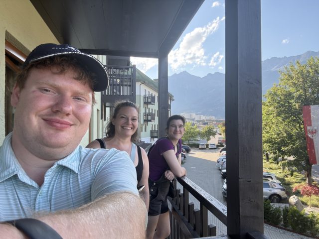 Selfie of Elliot in a cap with two friends on a wooden alpine balcony, mountains and a village street below.