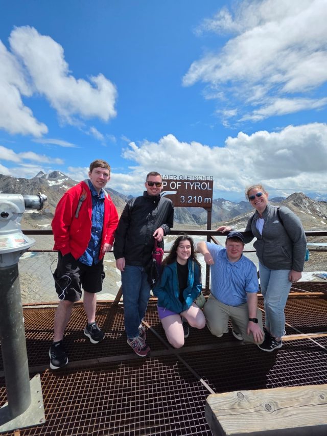 Elliot kneeling with four friends on a mesh viewing platform beneath a sign reading "Top of Tyrol 3,210 m", snow-streaked peaks and clouds behind them.