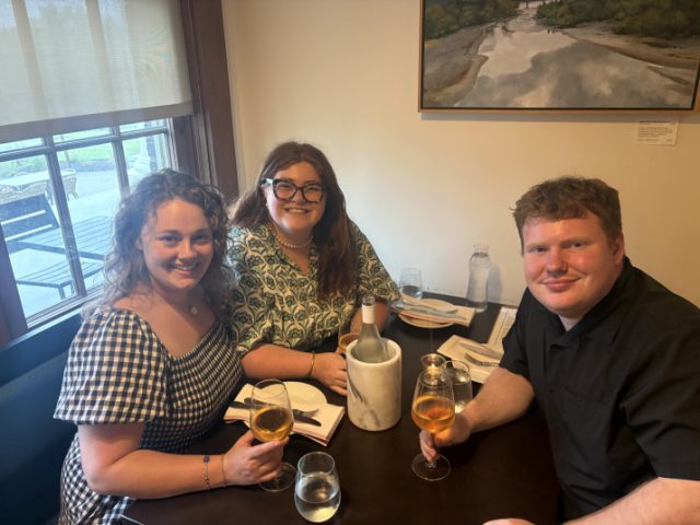 Elliot at a restaurant table with two friends, all three raising glasses of white wine toward the camera.