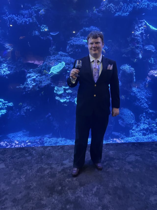 Elliot in a dark suit and patterned tie raising a glass in front of a vast blue coral reef aquarium window.